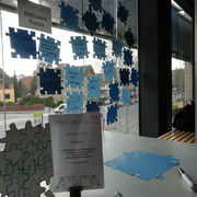 Zwei Schilder stehen auf einem Tisch, daneben liegen Puzzleteile aus blauem Papier. An einer Scheibe dahinter hängt ein Schild "Netzwerkpuzzle" und darum sind beschriftete, blaue Puzzleteile aufgehängt.